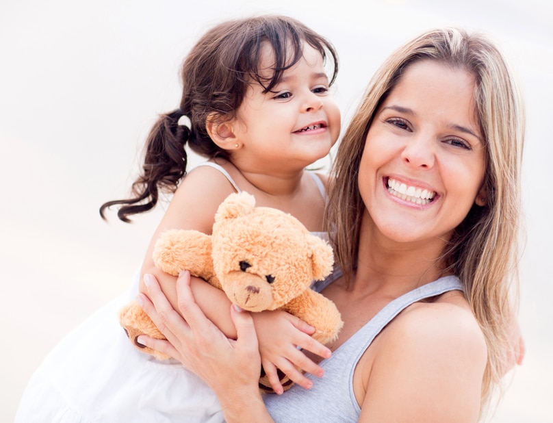 White mother carrying toddler daughter with a teddy bear and smiling at the camera