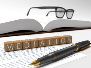 Wooden blocks with white letters on them spelling out mediation. The blocks sit on top of an unlined piece of paper with writing on it while a calligraphy pen sits in front of the blocks. A big book with square framed glasses on top is in the background.