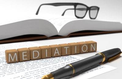 Wooden blocks with white letters on them spelling out mediation. The blocks sit on top of an unlined piece of paper with writing on it while a calligraphy pen sits in front of the blocks. A big book with square framed glasses on top is in the background.